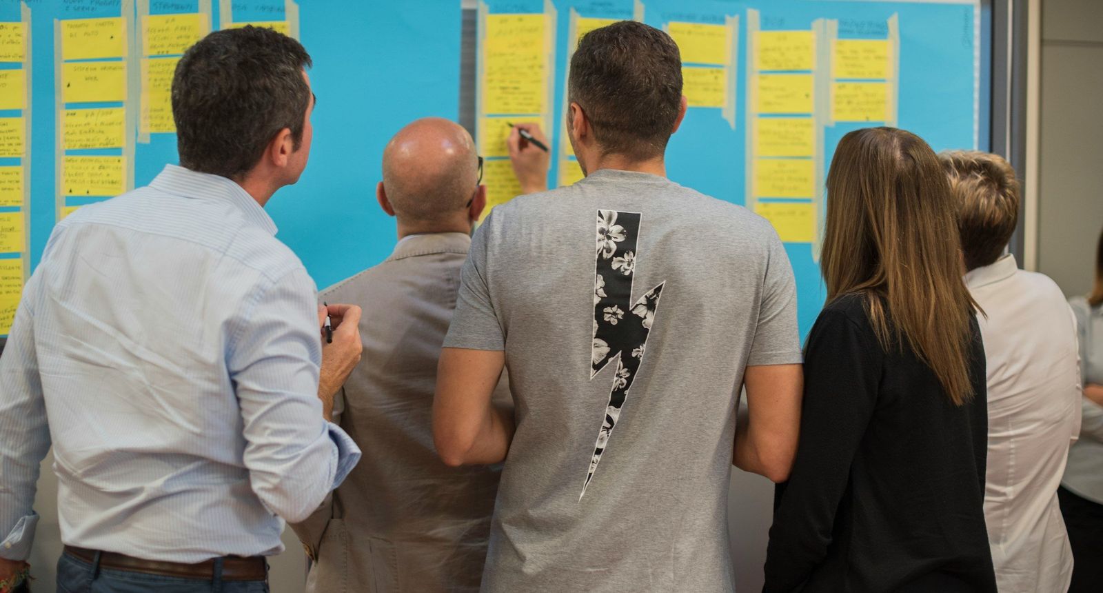 Five people seen from behind, standing at a wall of paper sheets covered in yellow sticky notes during a planning workshop.